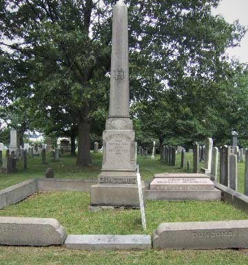 Tall granite obelisk in a cemetery, surrounded by other gravestones, green grass visible