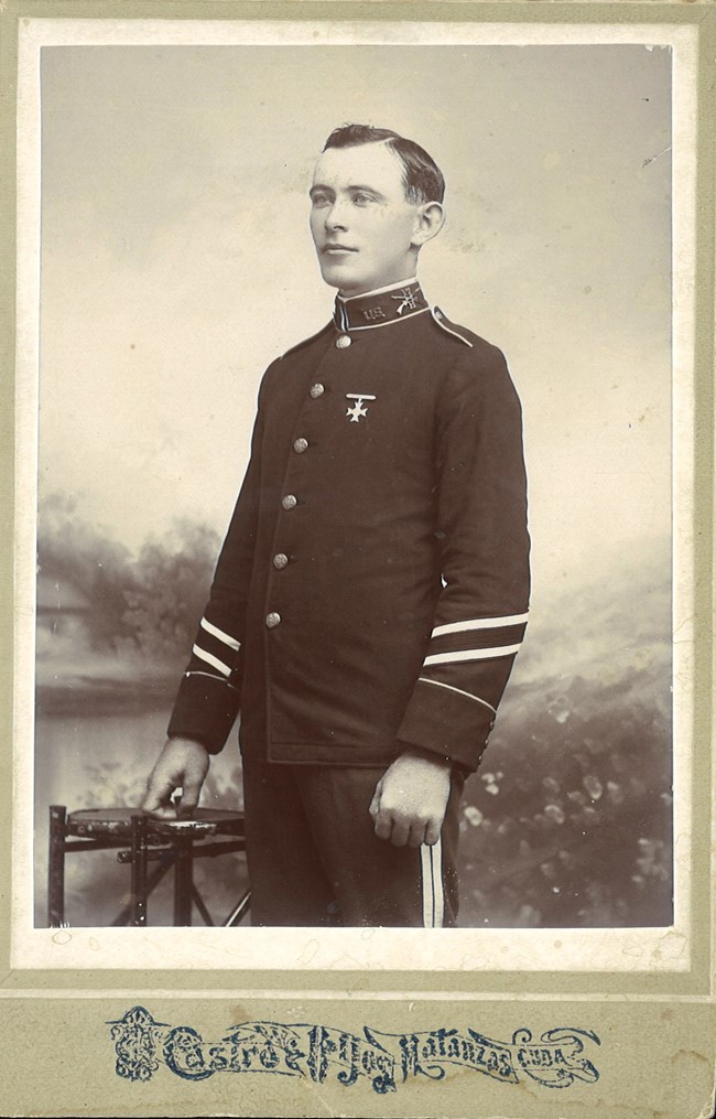 A black and white photo printed on a card. The photo shows a clean-shaven man wearing a military uniform posing in a photo studio.