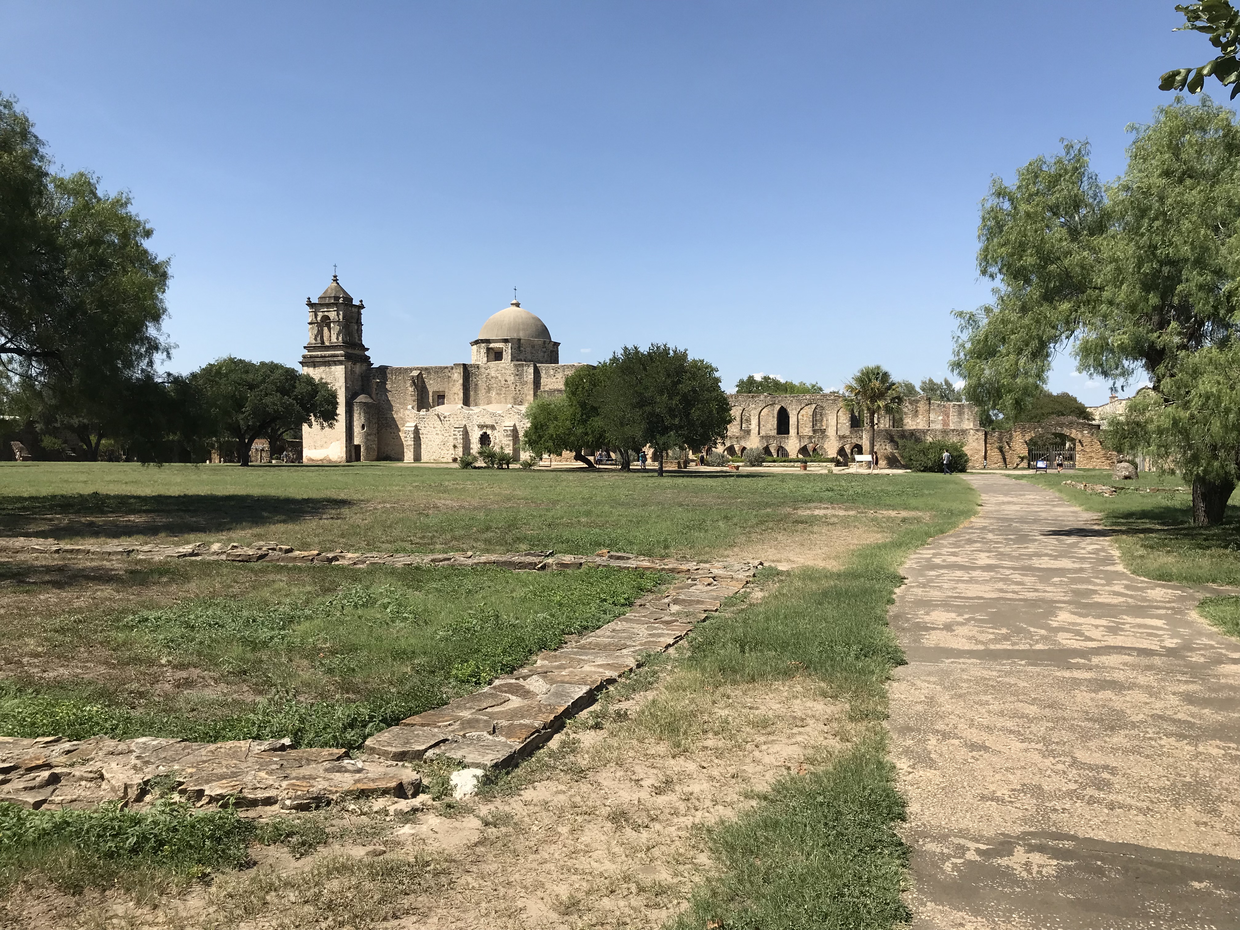 A green field and path leads to a distant spanish colonial mission.