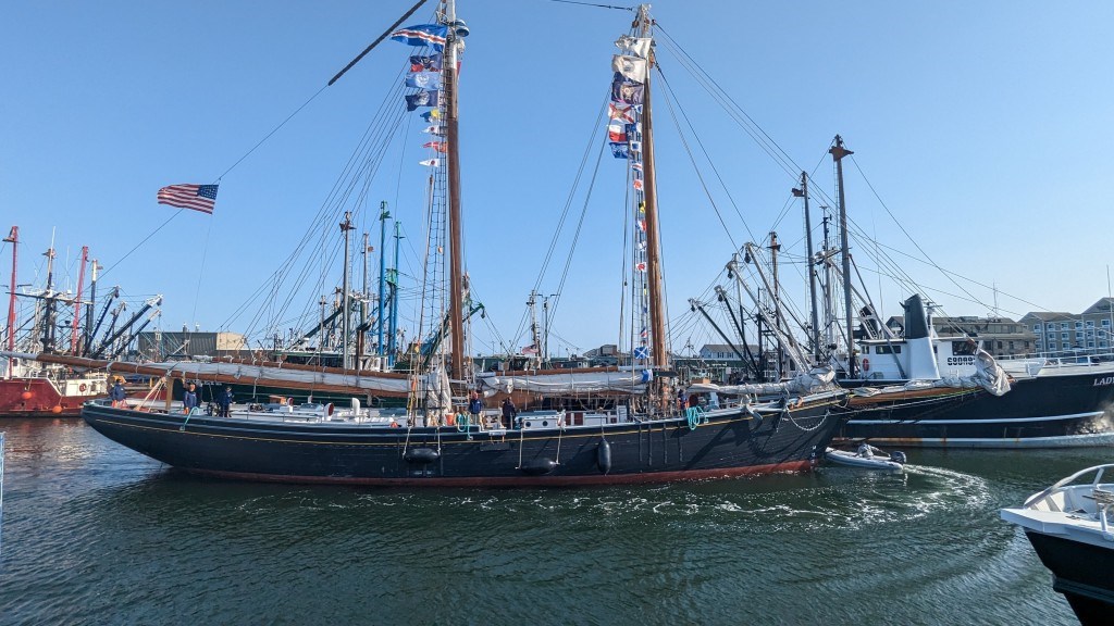 Schooner with two towering masts and sails lowered in a port surrounded by other vessels.