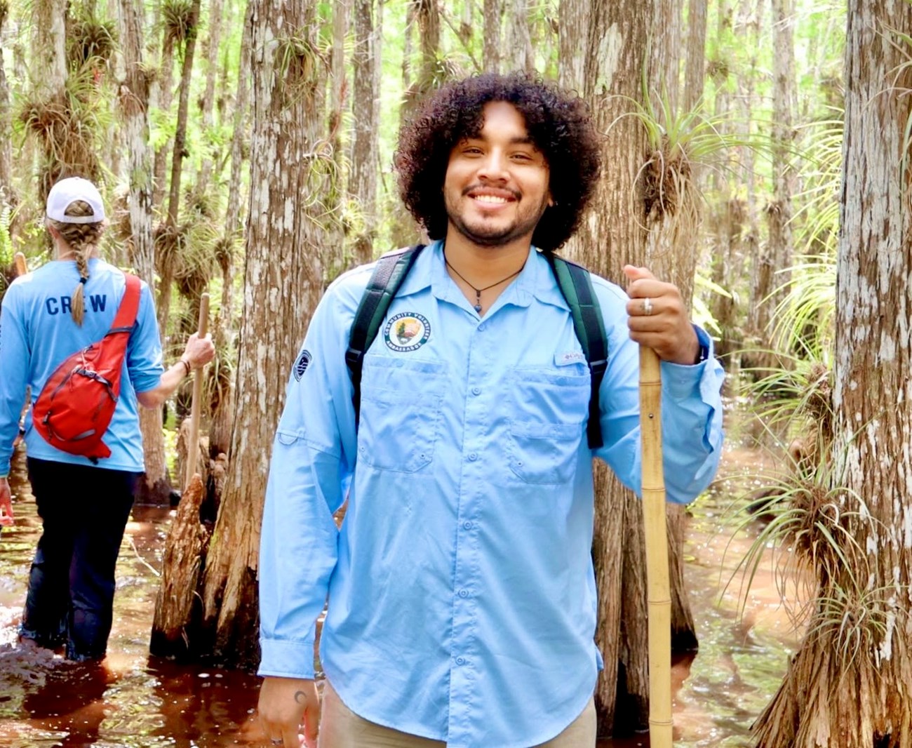 A young man smiles in front of a collection of tall trees