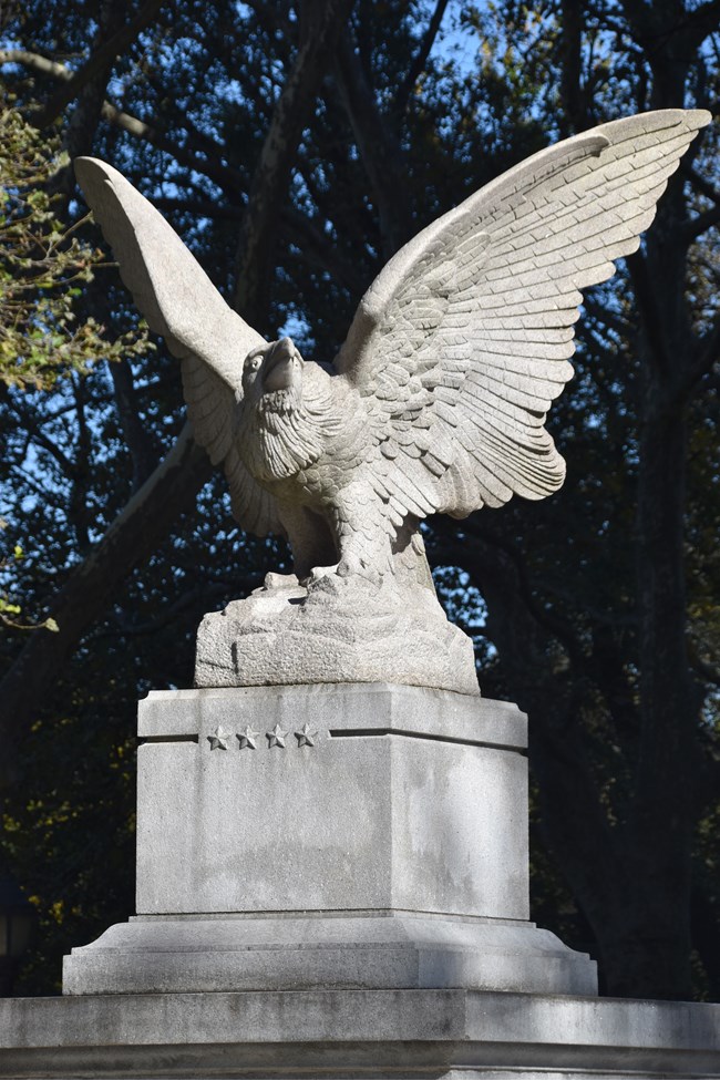 Image of a white granite eagle on a pedestal with trees in the background
