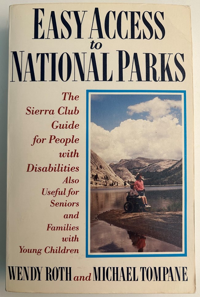 Book cover of Easy Access to National Parks: A Sierra Club Guide for People with Disabilities. Photo on cover of woman in wheel chair with man standing beside chair. Both are on edge of lake looking back at mountains.
