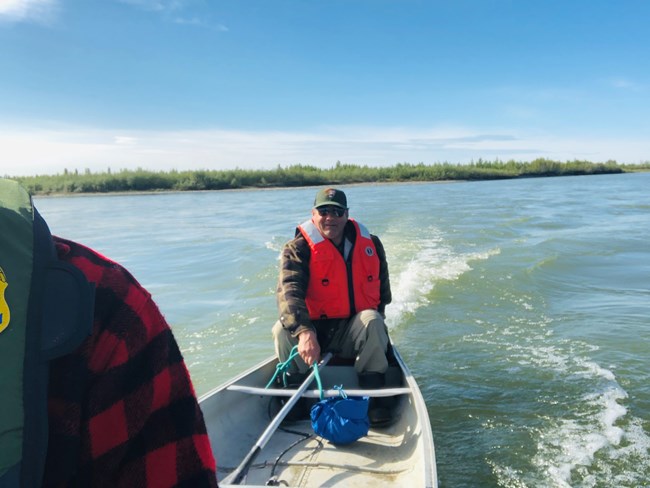Ed wears a life vest and stands in a canoe on a lake on a sunny day.