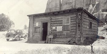 A small log cabin is seen at the base of a mountain.  A car is parked to the left of the cabin.