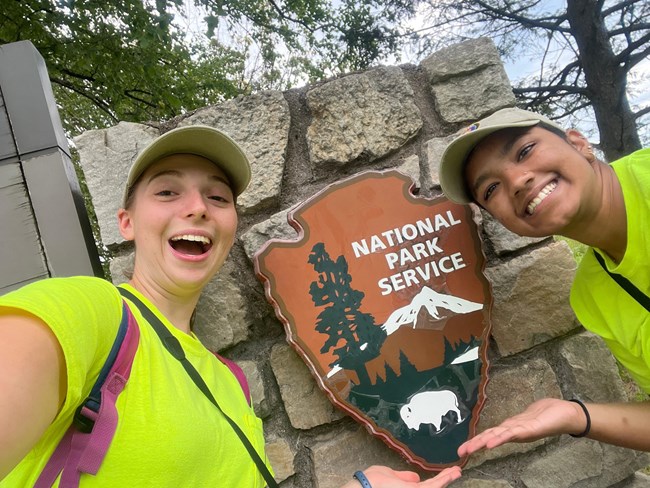 a light skinned girl and a dark skinned girl smile bigly wearing bright work shirts next to the national park service arrowhead