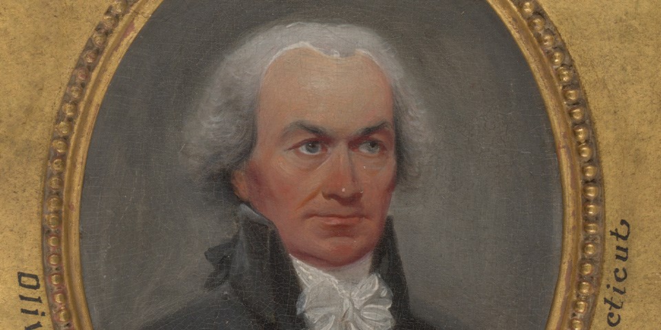 Ellsworth in black coat with high collar facing front-right.