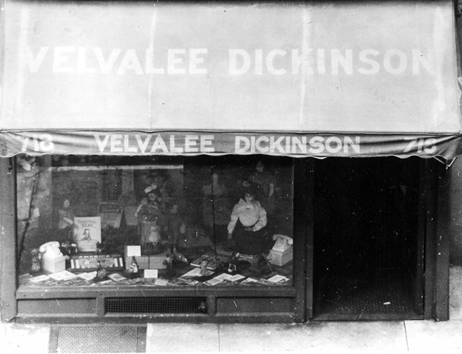 Black and white photo of a storefront. The awning reads “Velvalee Dickinson.” In the window are several dolls and accessories.