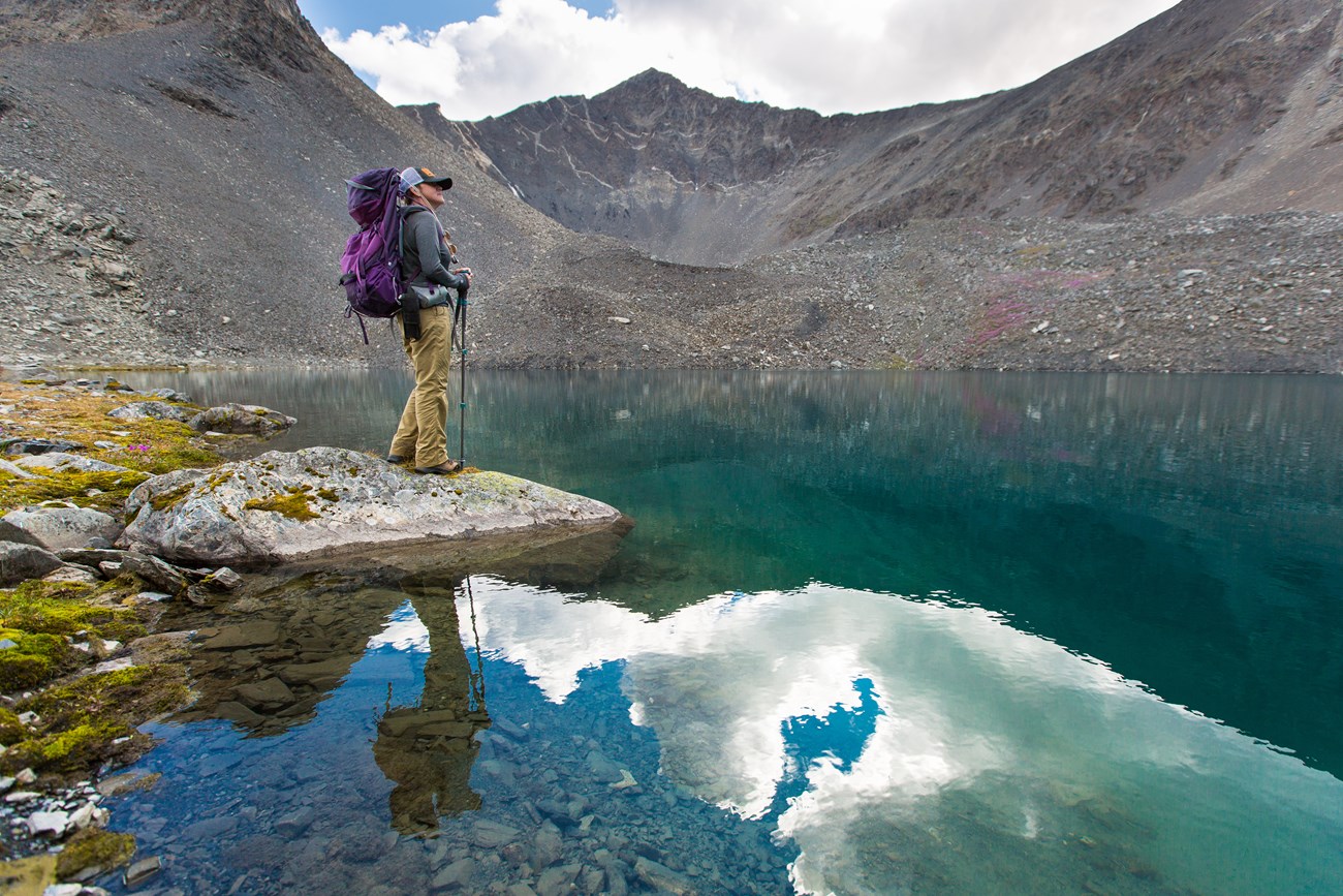 Hiker with purple backpack stands on the edge of a small blue green lake with jagged grey peaks in the background