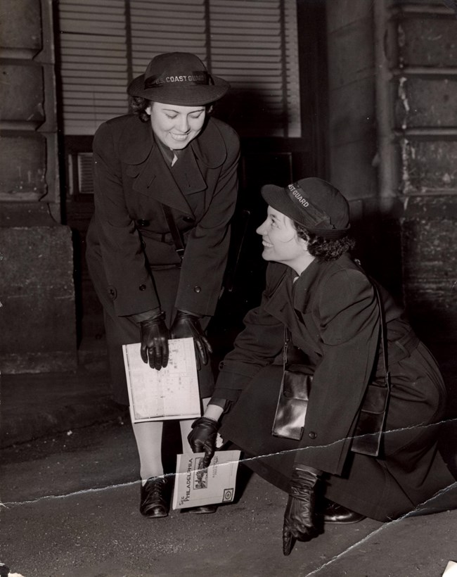 Black and white photo of two women in dark clothes, one kneeling and pointing, the other looking over her shoulder