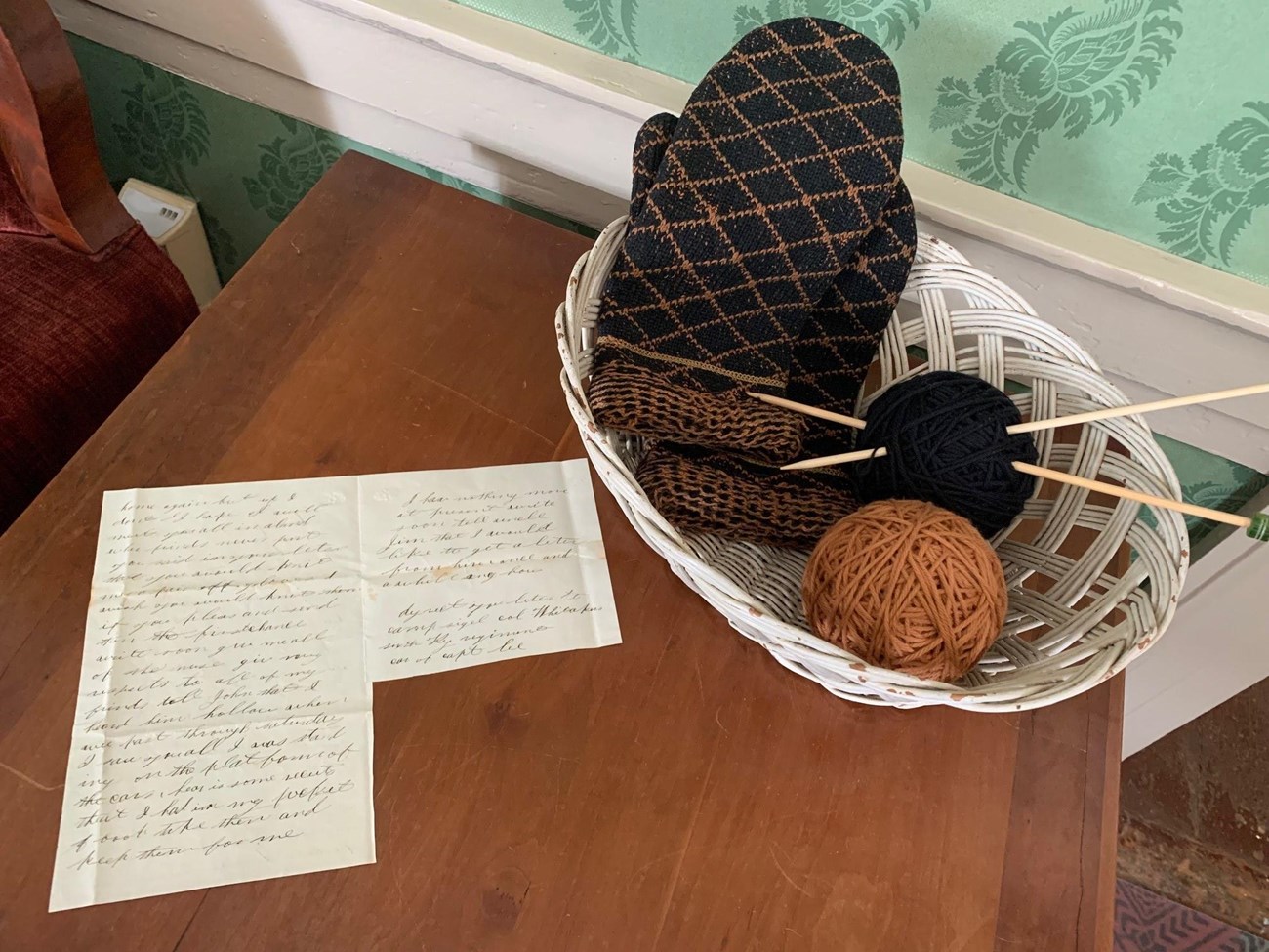 Handwritten letter sits on a table next to a white, wicker basket filled with black and tan yarn, knitting needles, and black and tan knit mittens.