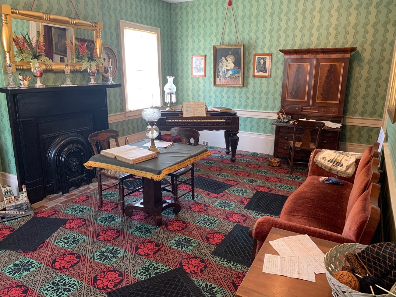 Color photograph of a Victorian Parlor. The walls are covered with a mint green wallpaper, stamped with darker green flora and fauna. Victorian era furnishings sit throughout the room.