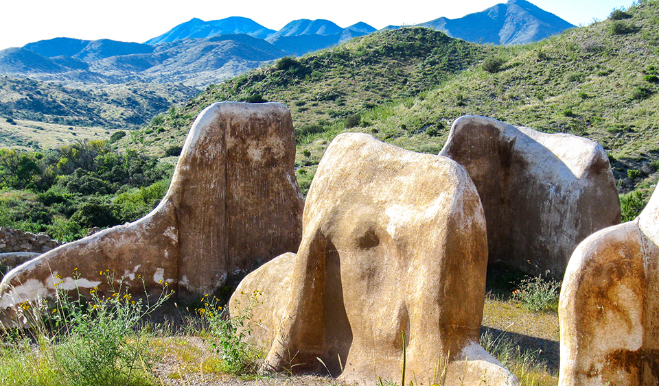 Ruins of several walls on a green, hilly landscape with brilliant blue mountains in the background.