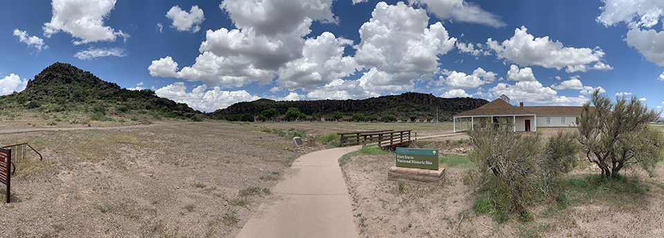 A landscape view of Fort Davis buildings, grassland, and rocky hills under fluffy clouds.