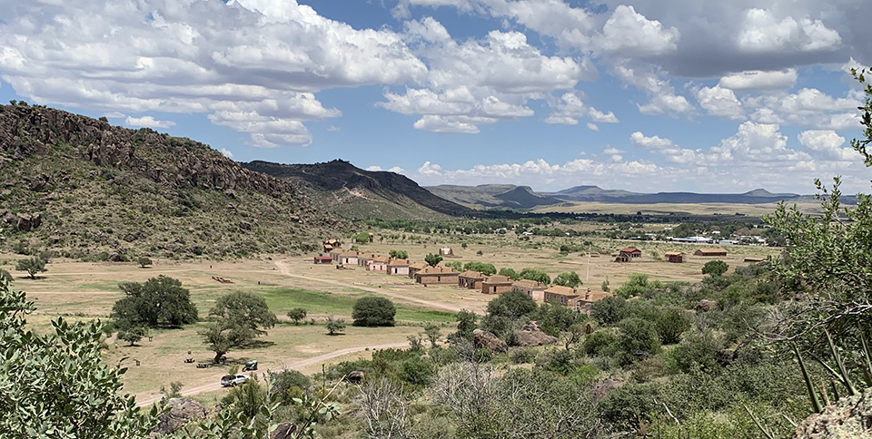 A landscape view of Fort Davis buildings, grassland, and rocky hills under fluffy clouds.