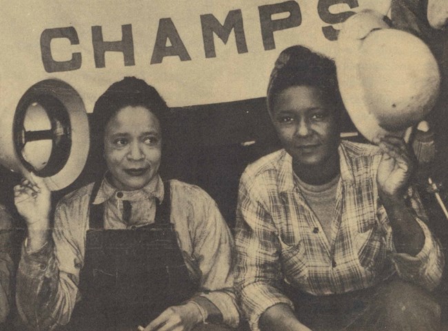 Two women of color wearing workers clothes hold up hard hats.