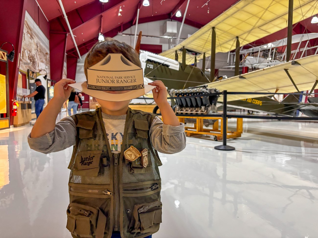 Kid wearing a Junior Ranger vest with a badge while holding a paper Junior Ranger hat over his face