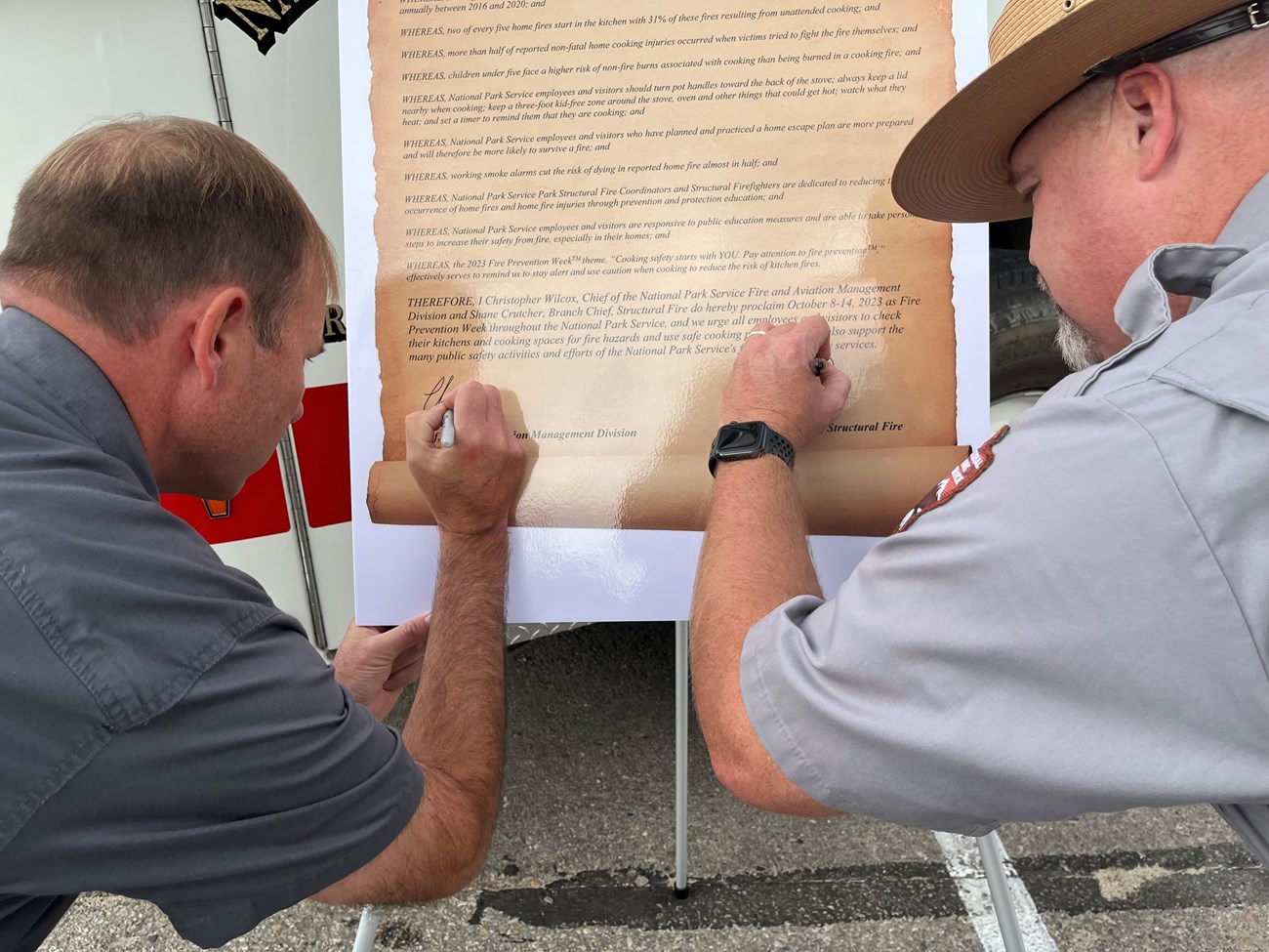 Two men sign a proclamation posted on an easel in front of a fire engine