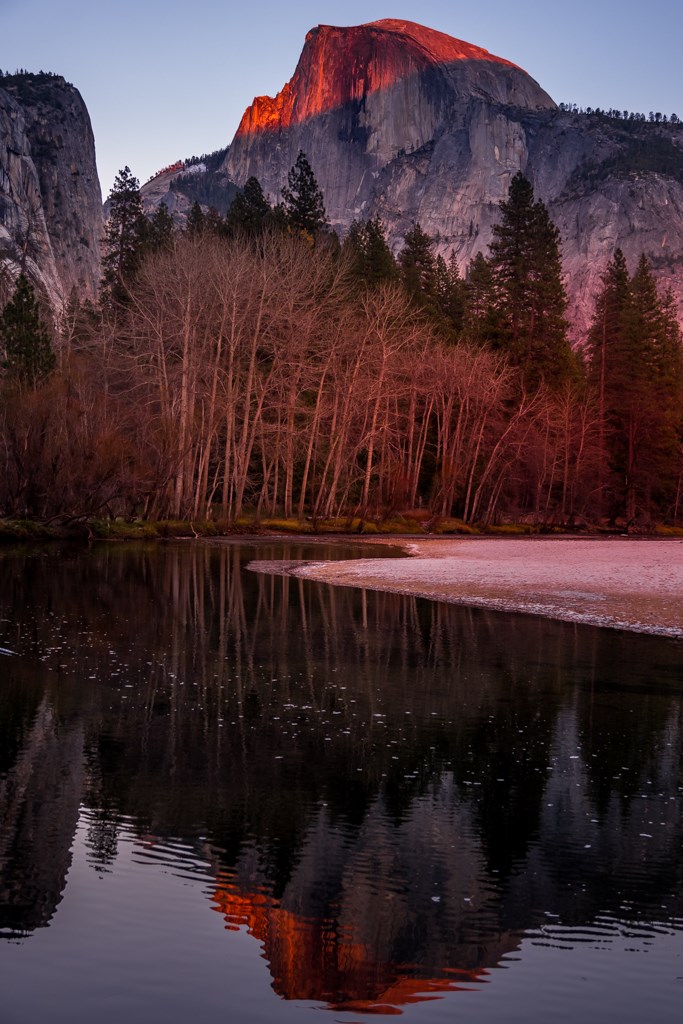 Photo of Half Dome in Yosemite National Park taken from Yosemite Valley during the fall sunset hour. The upper half of Half Dome's north face turns orange. Reflection of Half Dome in the Merced River.