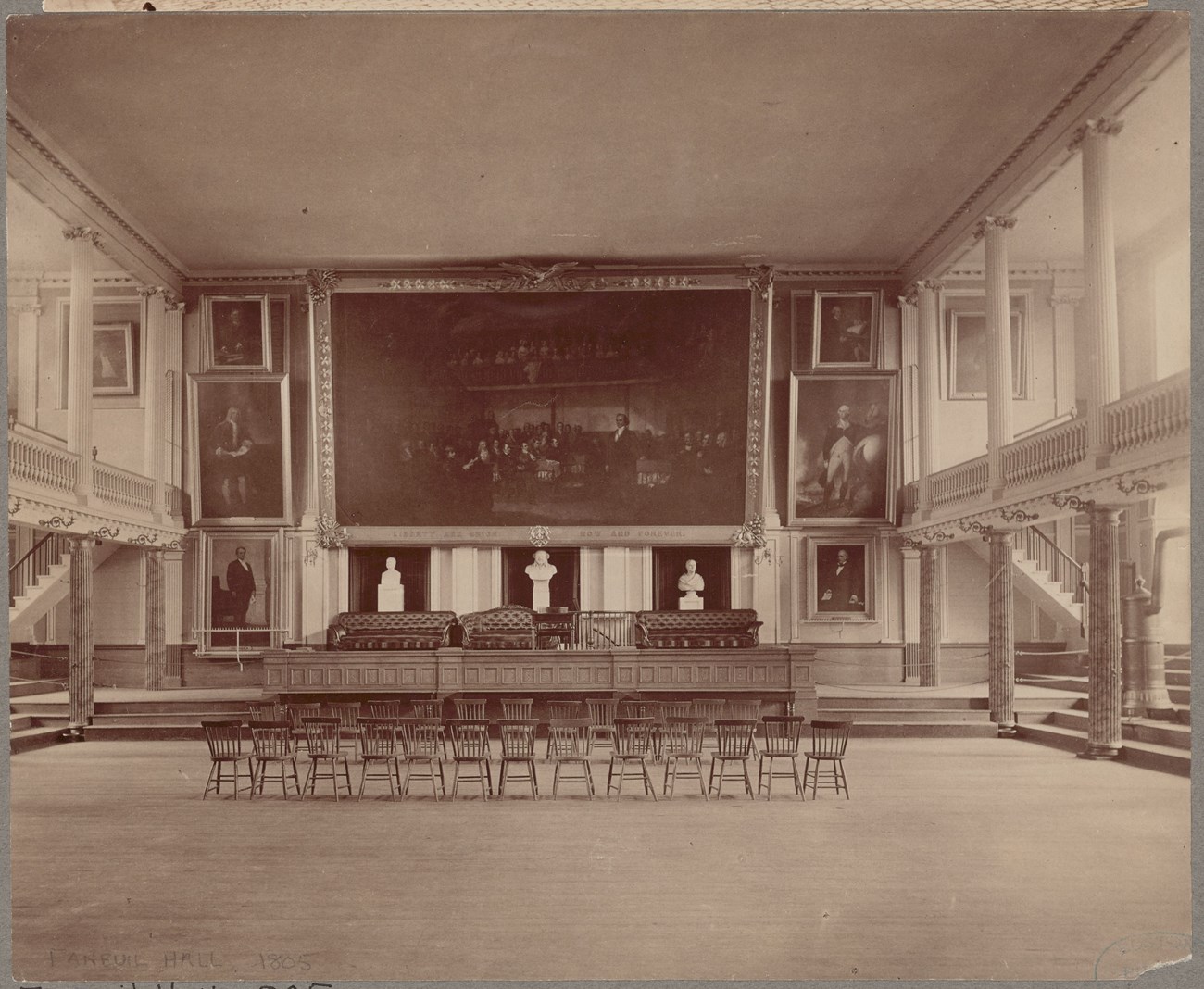 Sepia photo of the great hall of Faneuil Hall with a couple rows of chairs in front of the stage.