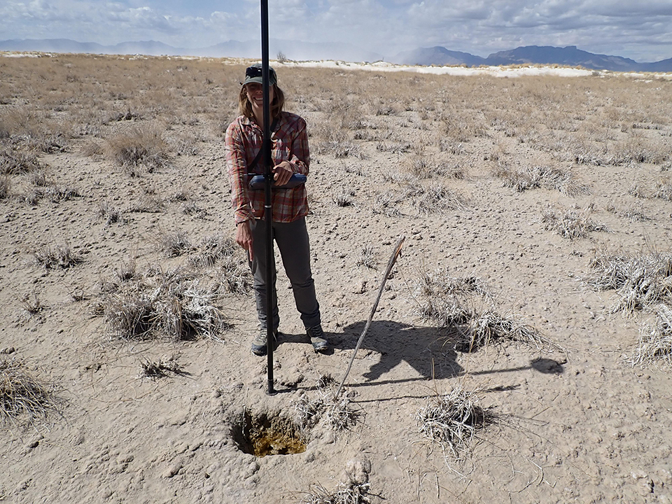 A person next to an upright pole pointing to a depression in a flat, white, sandy area with dispersed clumps of grass.