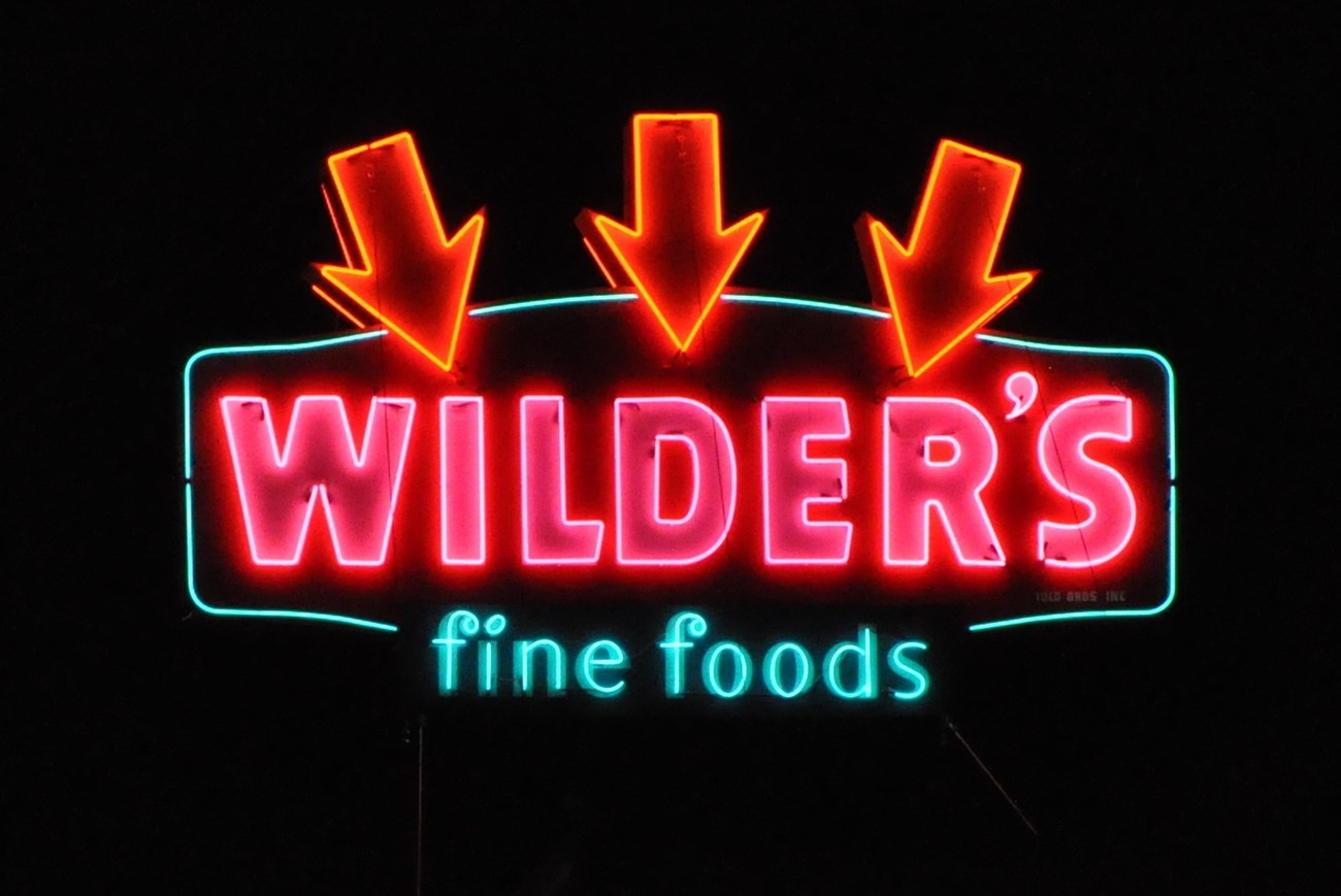 Three red neon arrows point to Wilder’s pink neon Fine Food in turquoise neon.