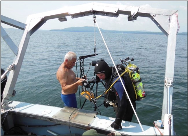 An unmasked diver reviews a site plan at the rear of the boat while another man checks equipment.