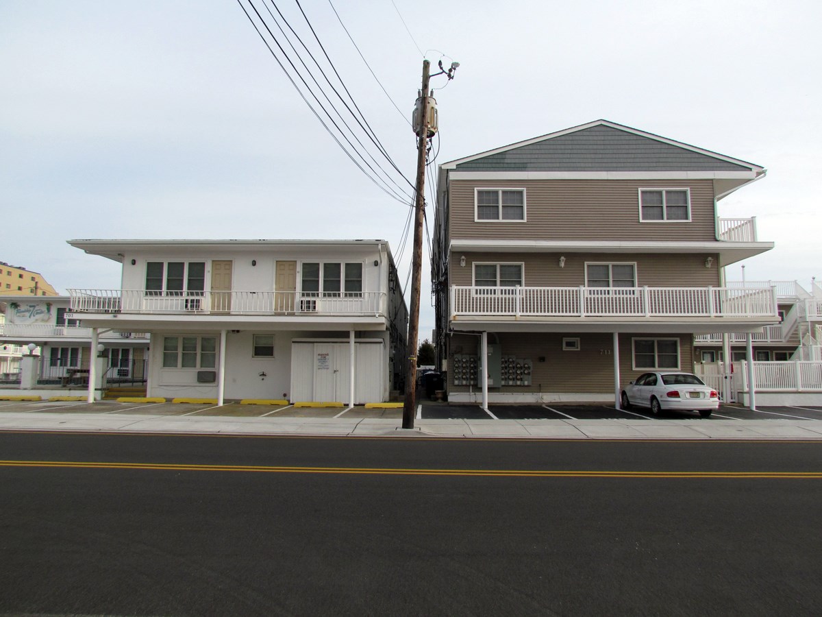 White two-story building with balcony next to tan three-story building with second floor balcony; parking in front.