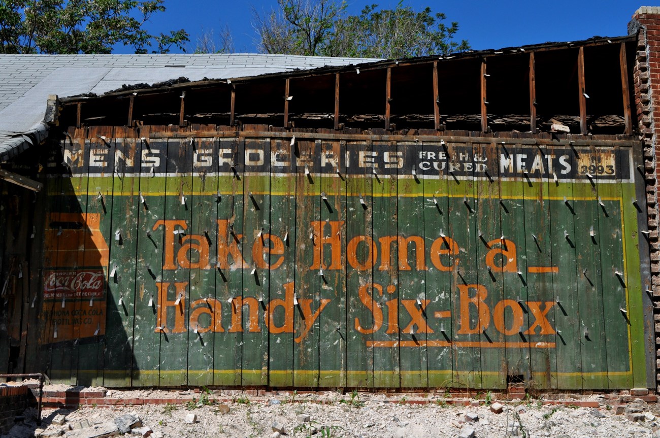 Weathered sign on a greenwood plank wall with Coca-Cola logo and orange text reading Take Home a handy six-box.