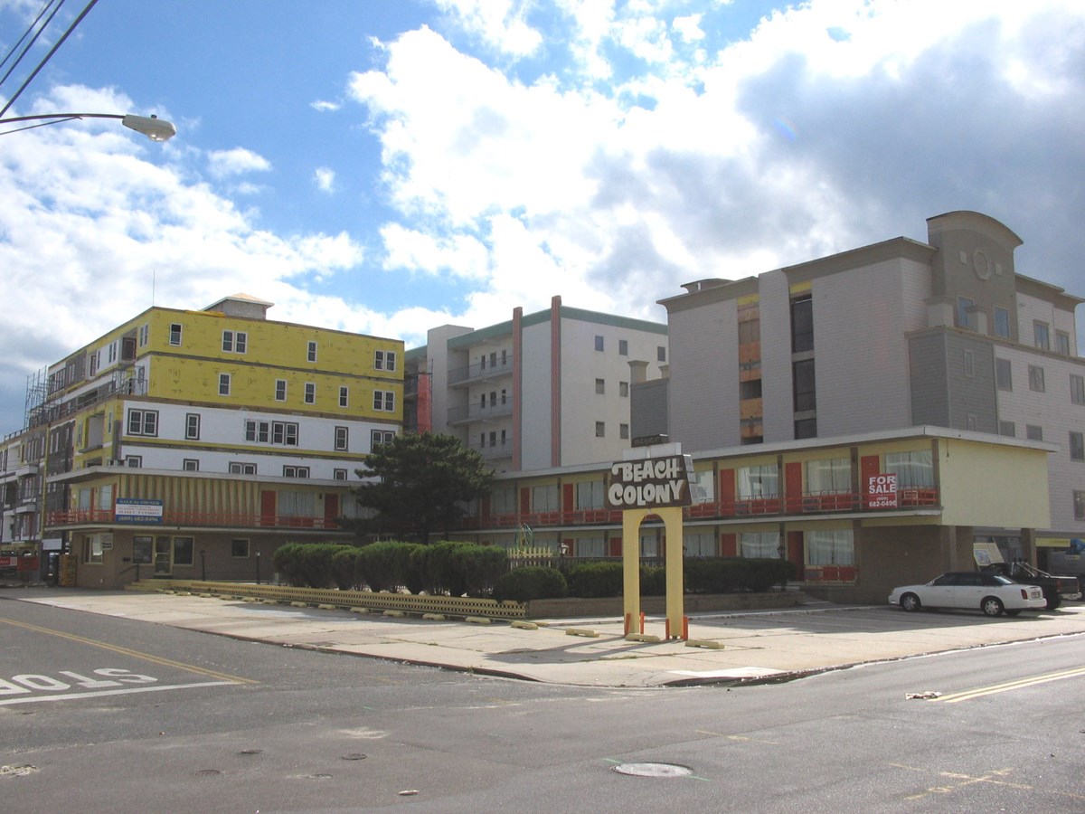 Two story weathered L shaped motel building in front of modern buildings with sign reading Beach Colony.