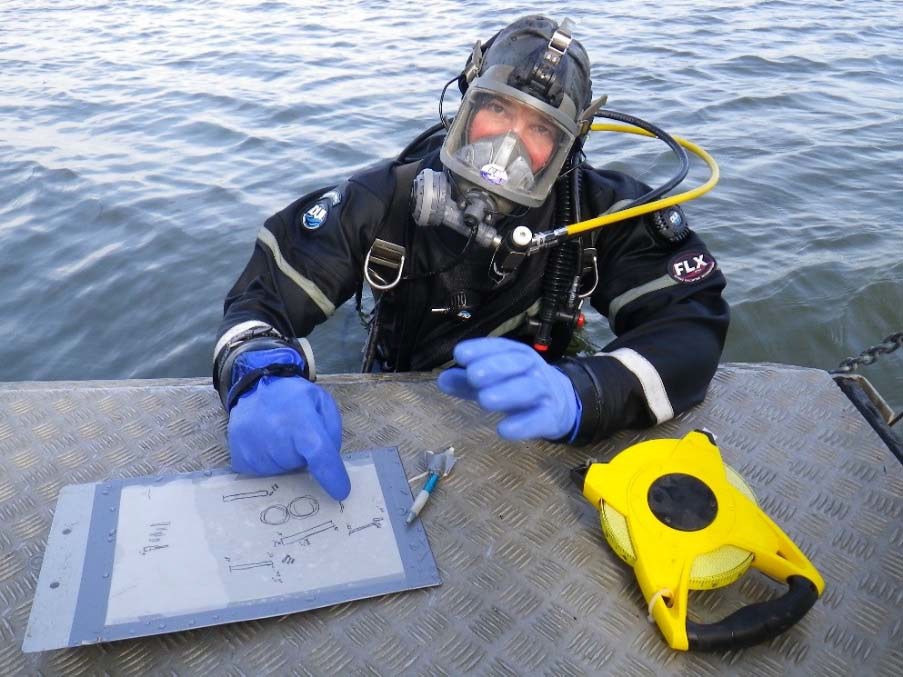 A diver with an Aga Mask and wireless comms sits partially submerged pointing to a sketchmap.