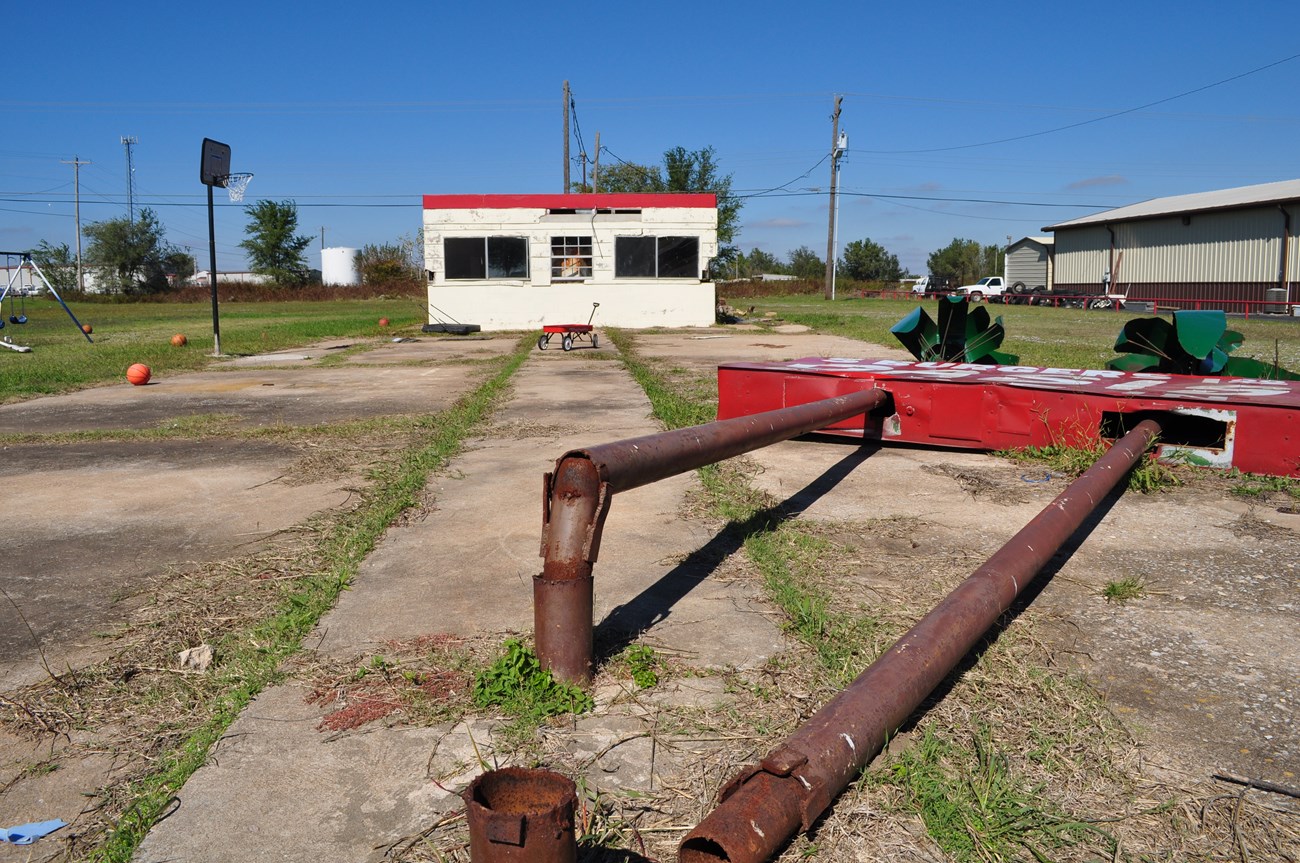 Grass growing in cement lot with small whit building in background. Foreground rusted poles of a collapsed sign.