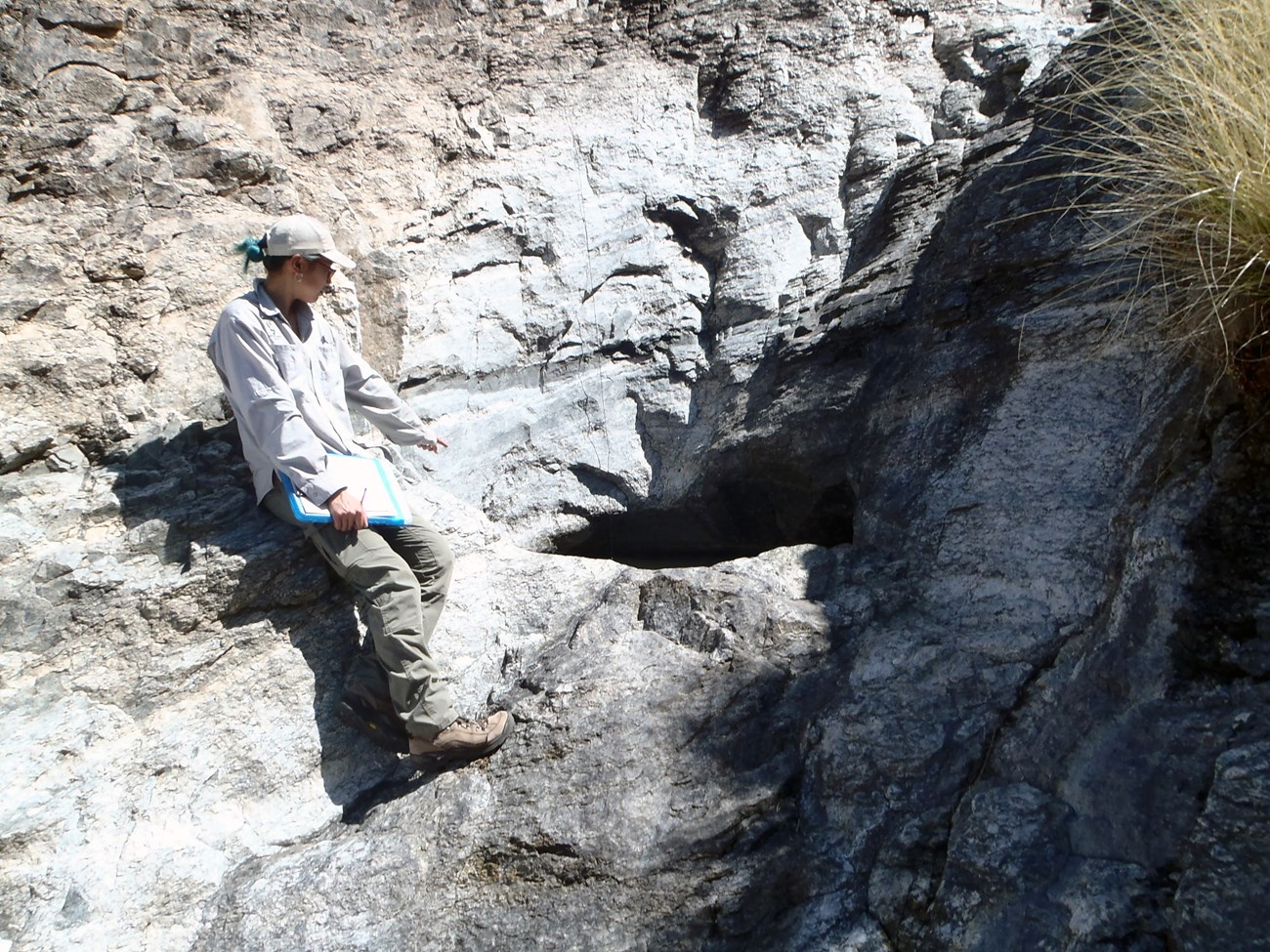 A person standing on a rock lip of a steep cliff next to a water filled depression in the rock of the cliff.