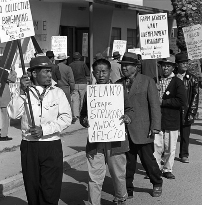 Several men walk in a line carrying AWOC picket signs in front of a building