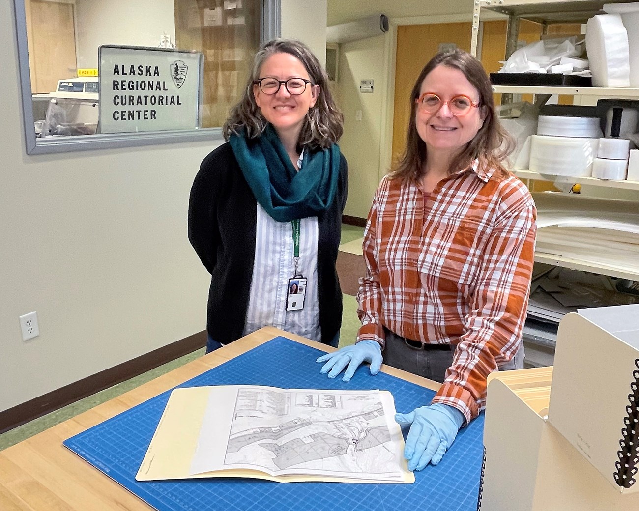 Two smiling people, one wearing gloves and handling a map in an open manila folder.