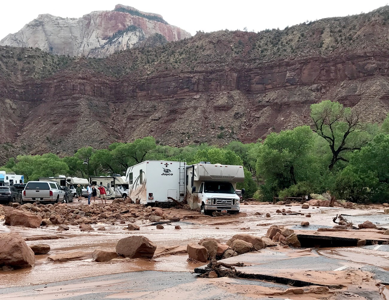 People standing around several RVs bogged down in the debris from a flood. Large rocks and mud are strewn about.