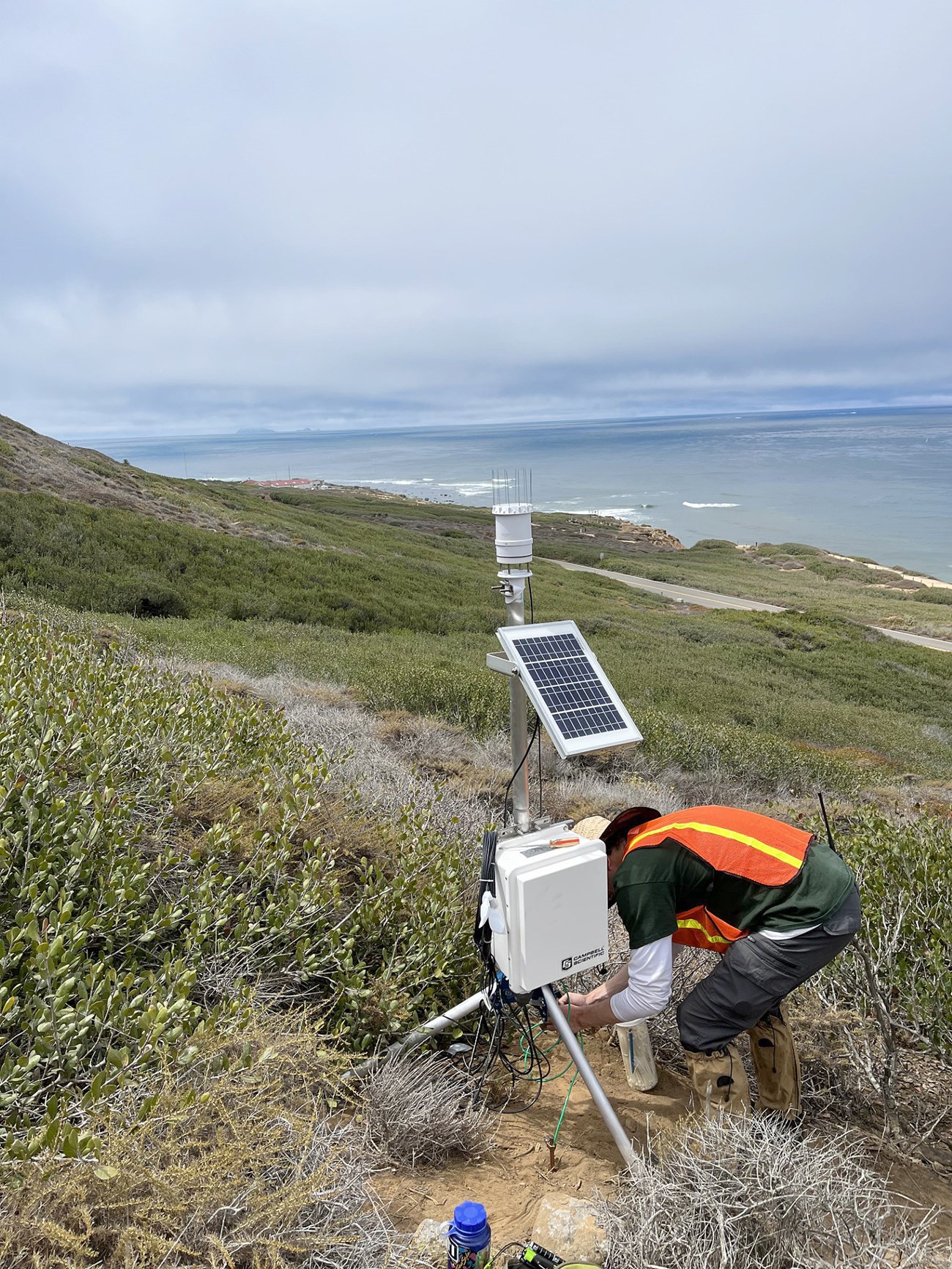 A man in an orange safety vest bends over while tending to a fog monitoring station. The Pacific Ocean is in the background.