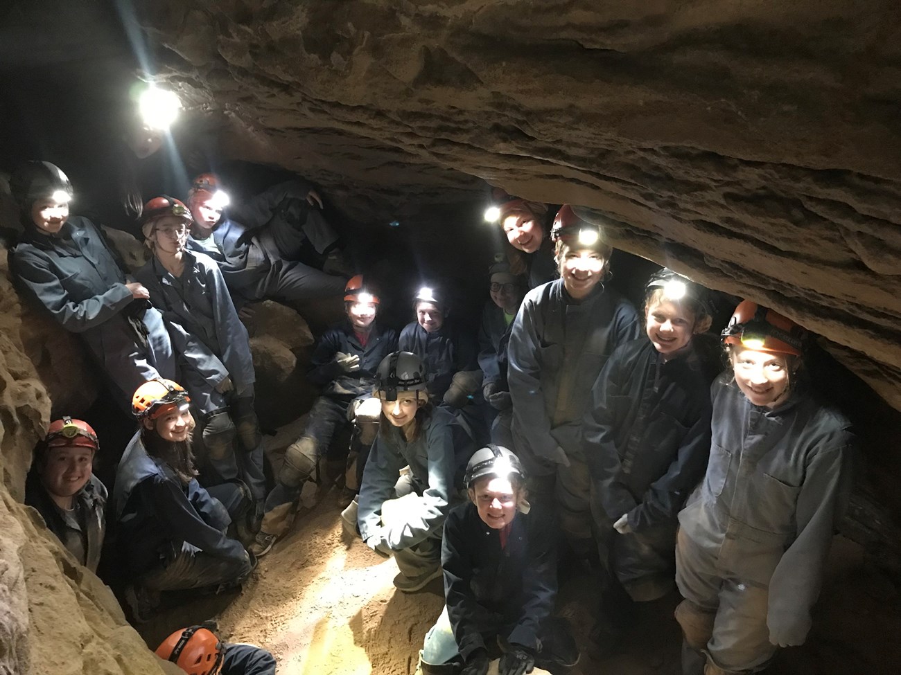 A group of young women wearing dark blue coveralls, helmets, and headlamps pose inside a small, dark, and rocky cave room.