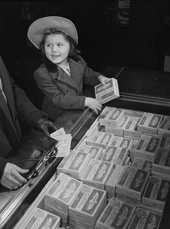Black and white photo of a smiling white girl wearing a hat and coat, holding a package of Birdseye frozen food. She is standing in front of a display case full of Birdseye product. We see only the white mother’s hands and purse.