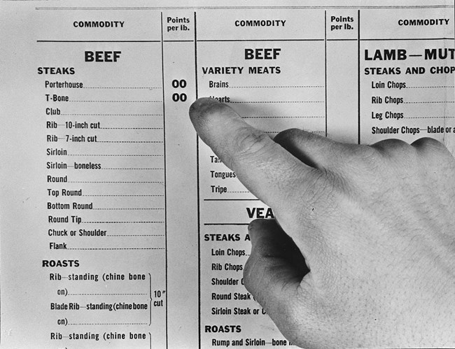 Black and white photo of a white hand pointing to an entry on a sample point rationing table. Only part of the table shows. There are separate entries for beef steaks, roasts, and variety meats; lamb steaks and chops; veal steaks and roasts.
