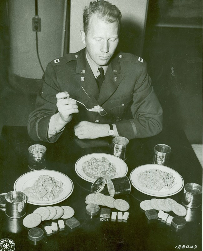 Black and white photo. The soldier is white. Three C-Ration meals are shown. Main courses are served on china plates, with extras laid out in front.