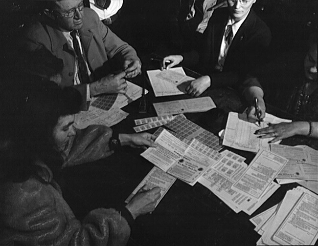 Black and white photo of white men and women sitting at a table. Some are filling out forms. The table surface is covered with paperwork and ration stamps.