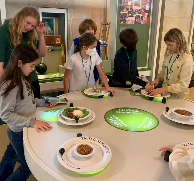 Children stand around a table while examining models of dishes and foods