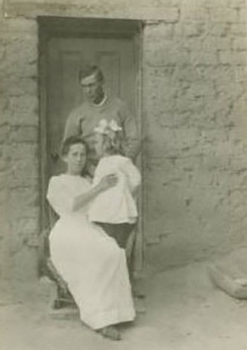 A man stands behind a woman holding a child. His arms are folded in front of him as looks down at smiling at his family.