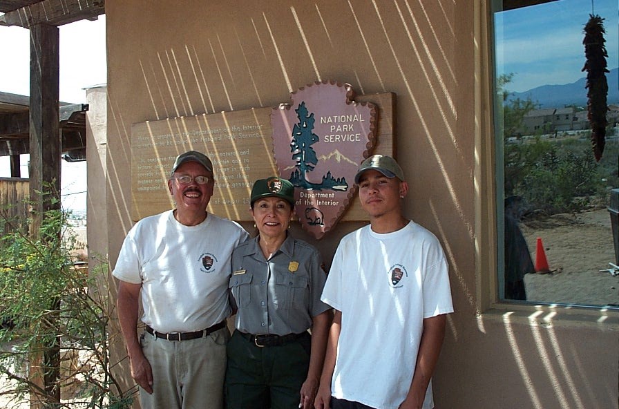 Judith Cordova in her NPS uniform poses in between a man and a teenager wearing white t-shirts. They stand in front of an NPS sign hung on an adobe building.