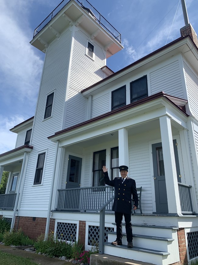 A man dressed in a black lighhouse keeper uniform waves on the front steps of a white lighthouse.