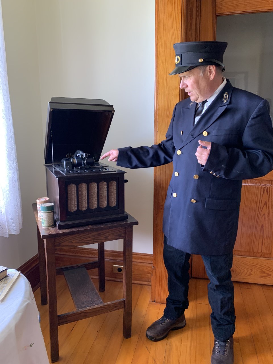 A man dressed in a black lighthouse keeper uniform pointing towards an Ambrola music machine.