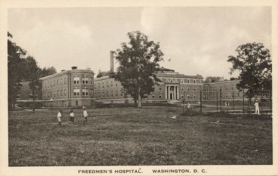 Sepia post card of a large three wing brick building with columns. Large trees and an open field with African American children walking through it.