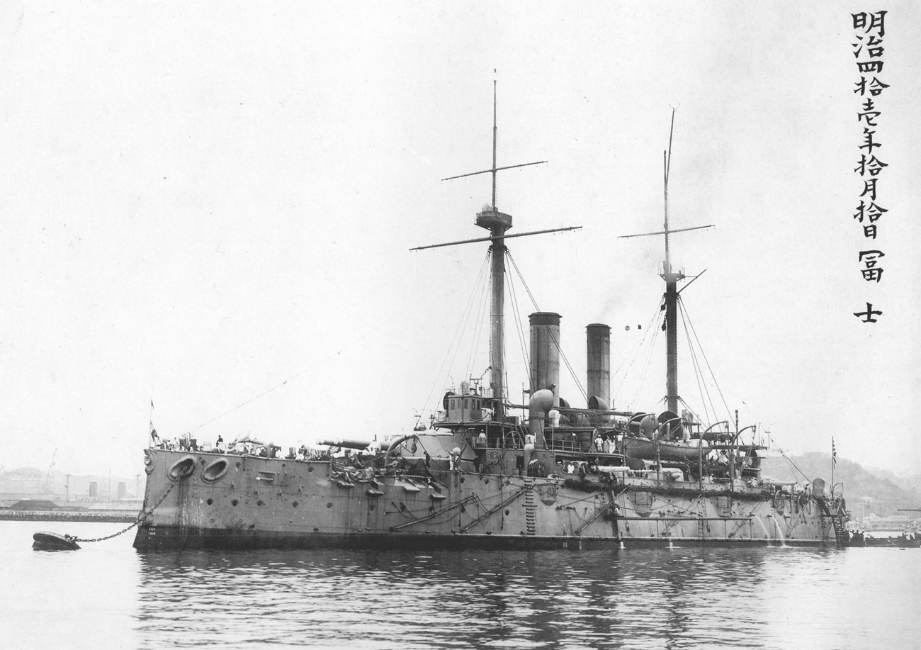 Black and white photo of large battleship with two smokestacks in the middle and two masts.