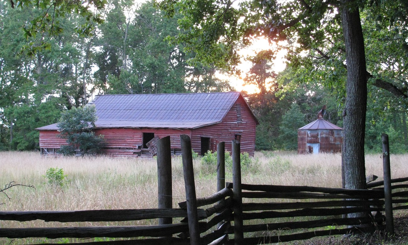 A fence stands in front of an old weathered red barn stands in a field at sunset.